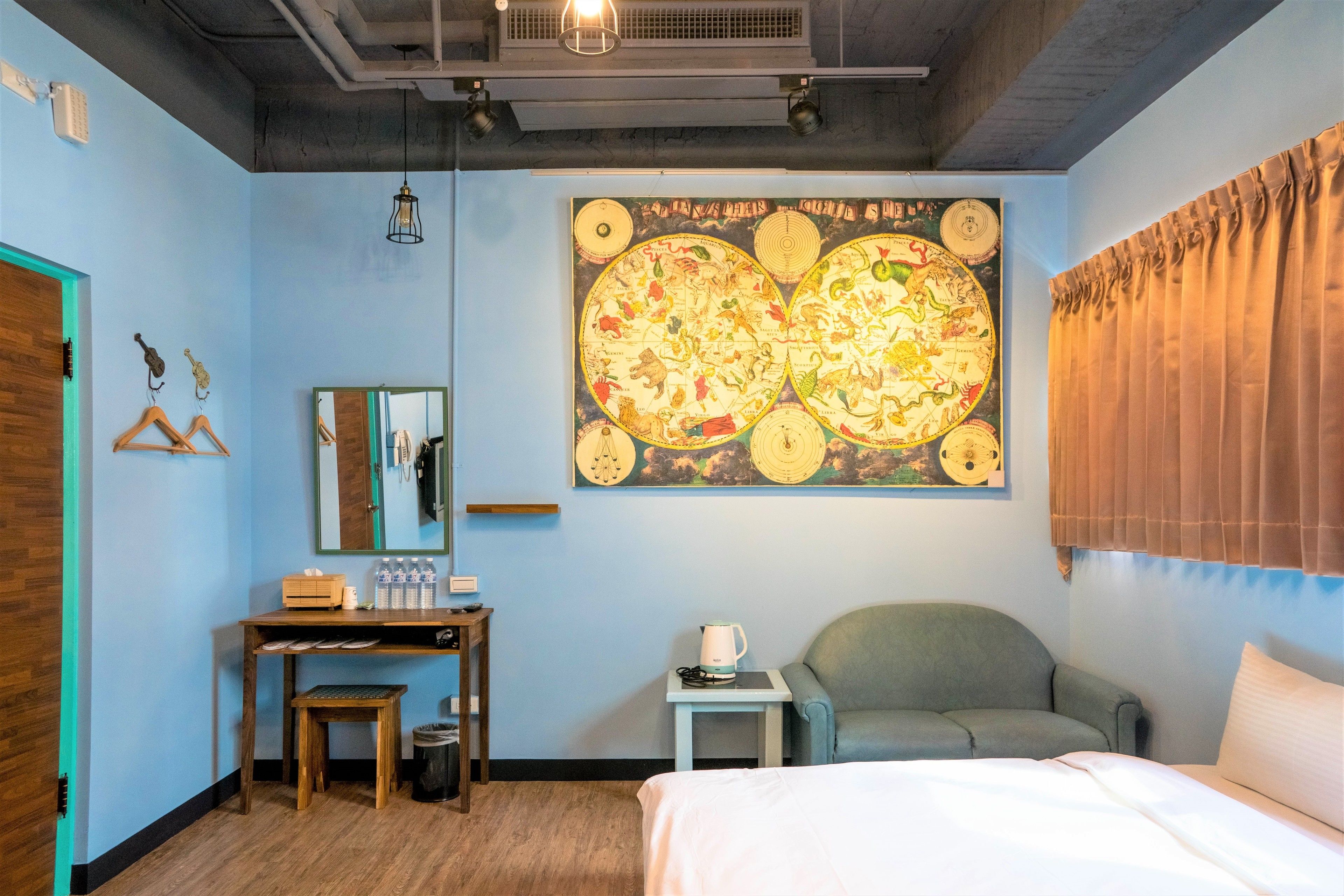 A hotel room with a large wall map, a desk with a mirror, a chair, a bed, and a small table with a kettle.