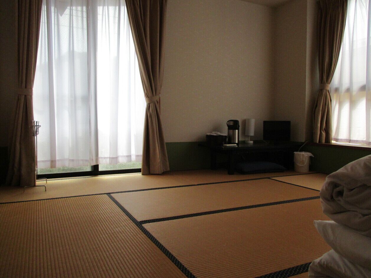A room with tatami flooring, a bed with a white blanket, a small table with a kettle, and a window with sheer curtains.