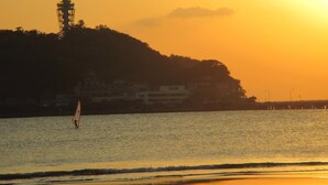 A sunset over a body of water with a sailboat, a lighthouse, and buildings on a hill. - Akitaya (Kamakura)