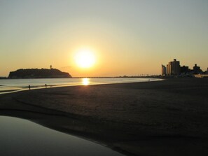 A beach with a sunset, calm water, and buildings in the background. - Akitaya (Kamakura)
