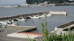 A marina with boats docked, a lighthouse, and a hill with buildings in the background. - Akitaya (Kamakura)