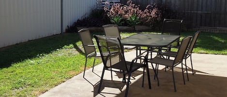 A patio with a table and chairs, surrounded by a white fence and greenery.