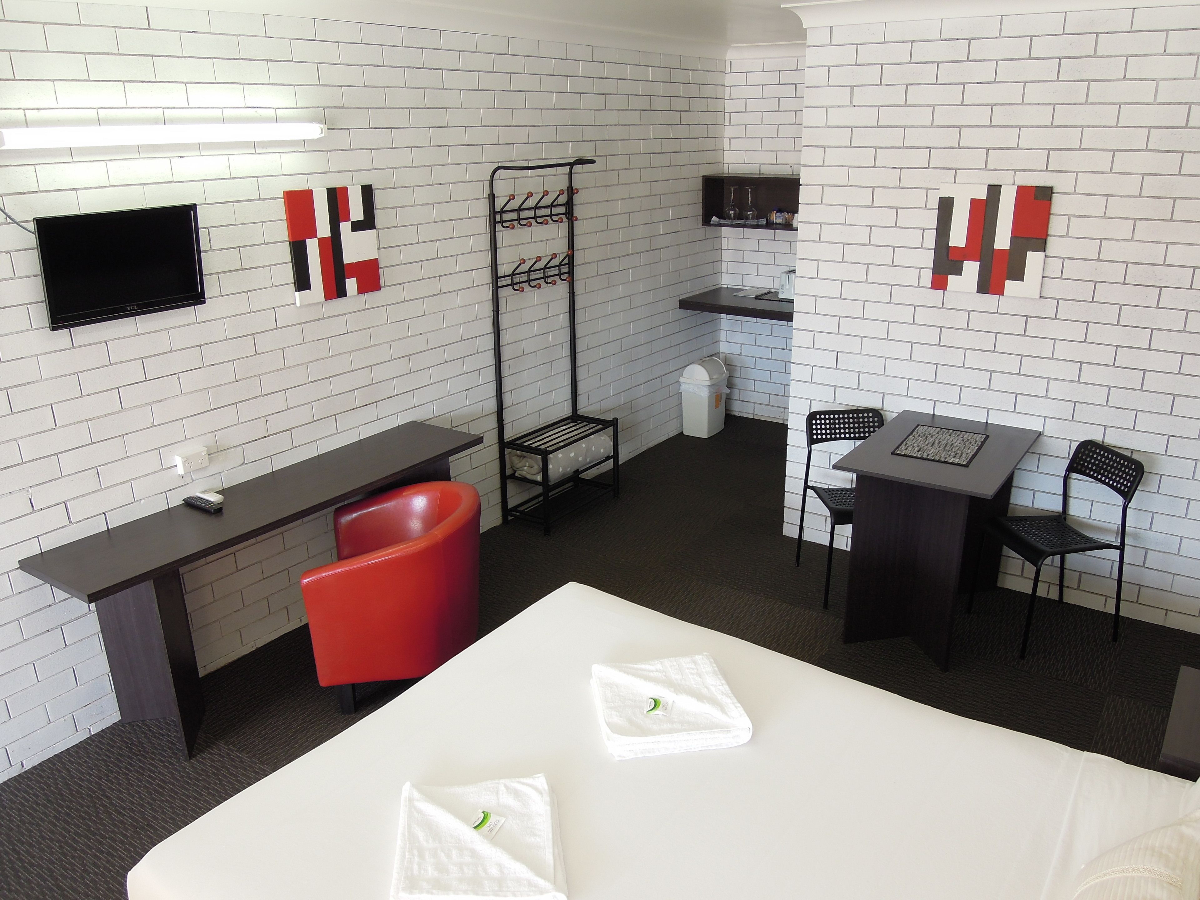 A hotel room with a white brick wall, a flat-screen TV mounted above a desk, a red chair, a small table with a chair, and a bed with white linens.
