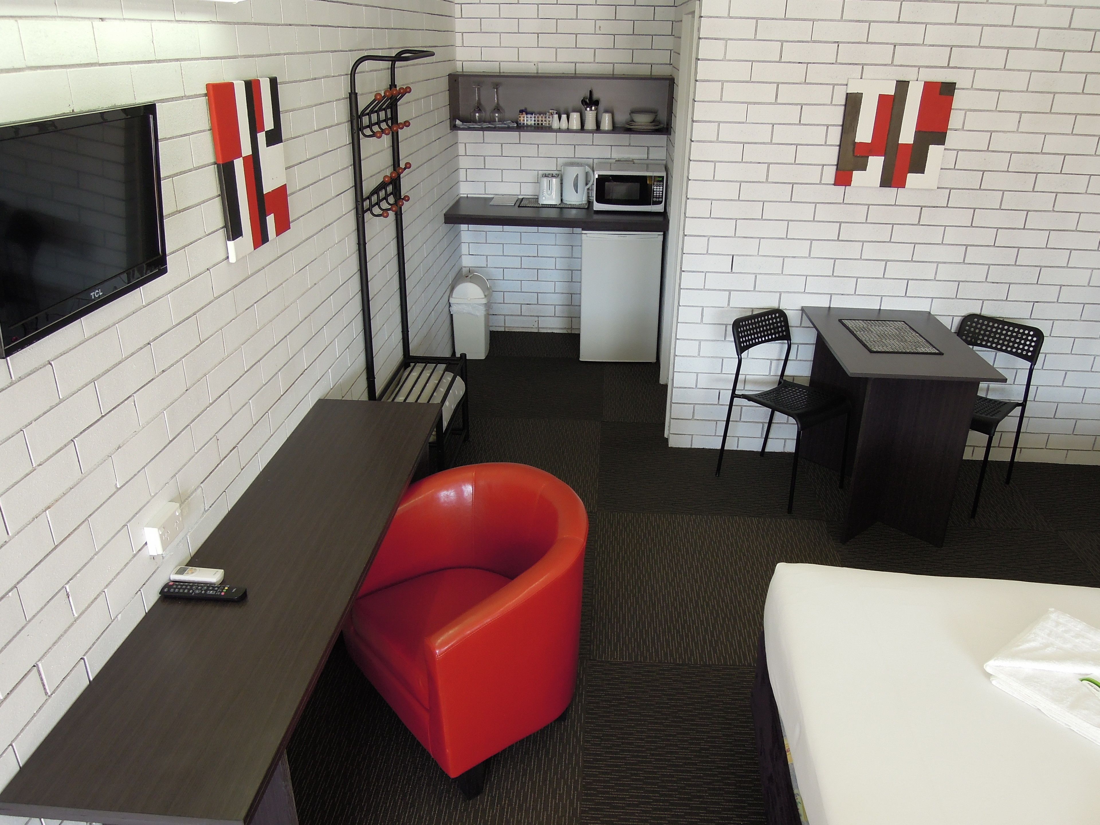 A compact hotel room with a flat-screen TV, a red chair, a desk, a small kitchenette, and a bed.