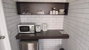 Una kitchenette compacta con microondas, tostadora y cafetera sobre una encimera oscura, un pequeño cubo de basura y un estante con vasos y platos.