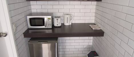 Una kitchenette compacta con microondas, tostadora y cafetera sobre una encimera oscura, un pequeño cubo de basura y un estante con vasos y platos.
