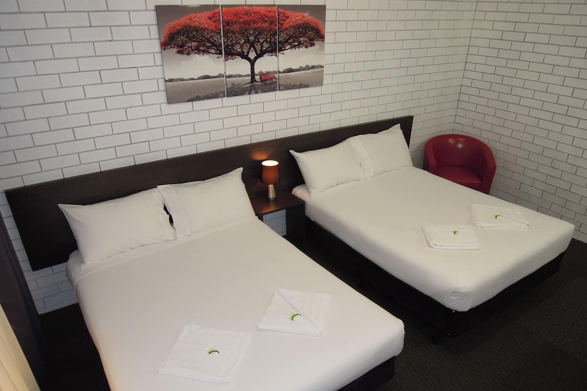 A hotel room with two beds, white linens, a red chair, a nightstand with a lamp, and a wall-mounted picture of a tree.