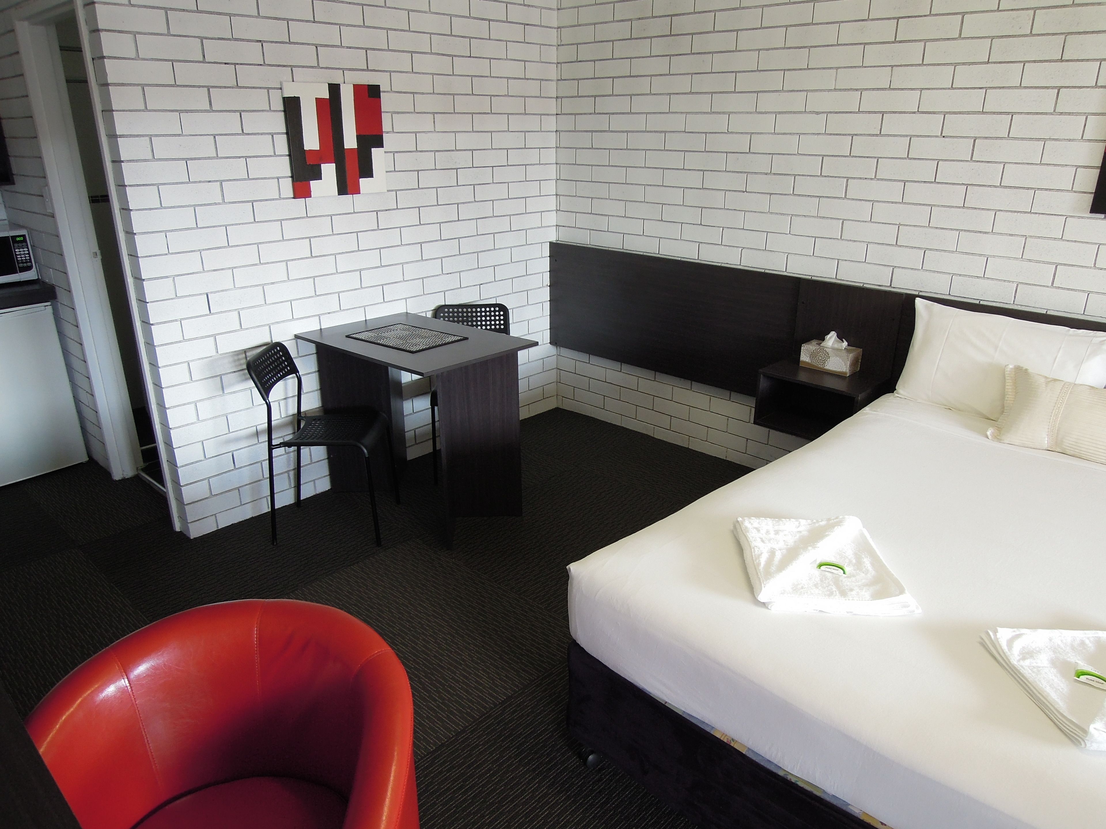 A hotel room with a white brick wall, a bed with white bedding, a desk with a chair, a red chair, and a TV on a stand.