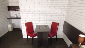 Una habitación de hotel pequeña con pared de ladrillo blanco, alfombra marrón oscuro, una cama individual, una pequeña mesa con dos sillas rojas, una mesita de noche con lámpara y un cesto de basura.