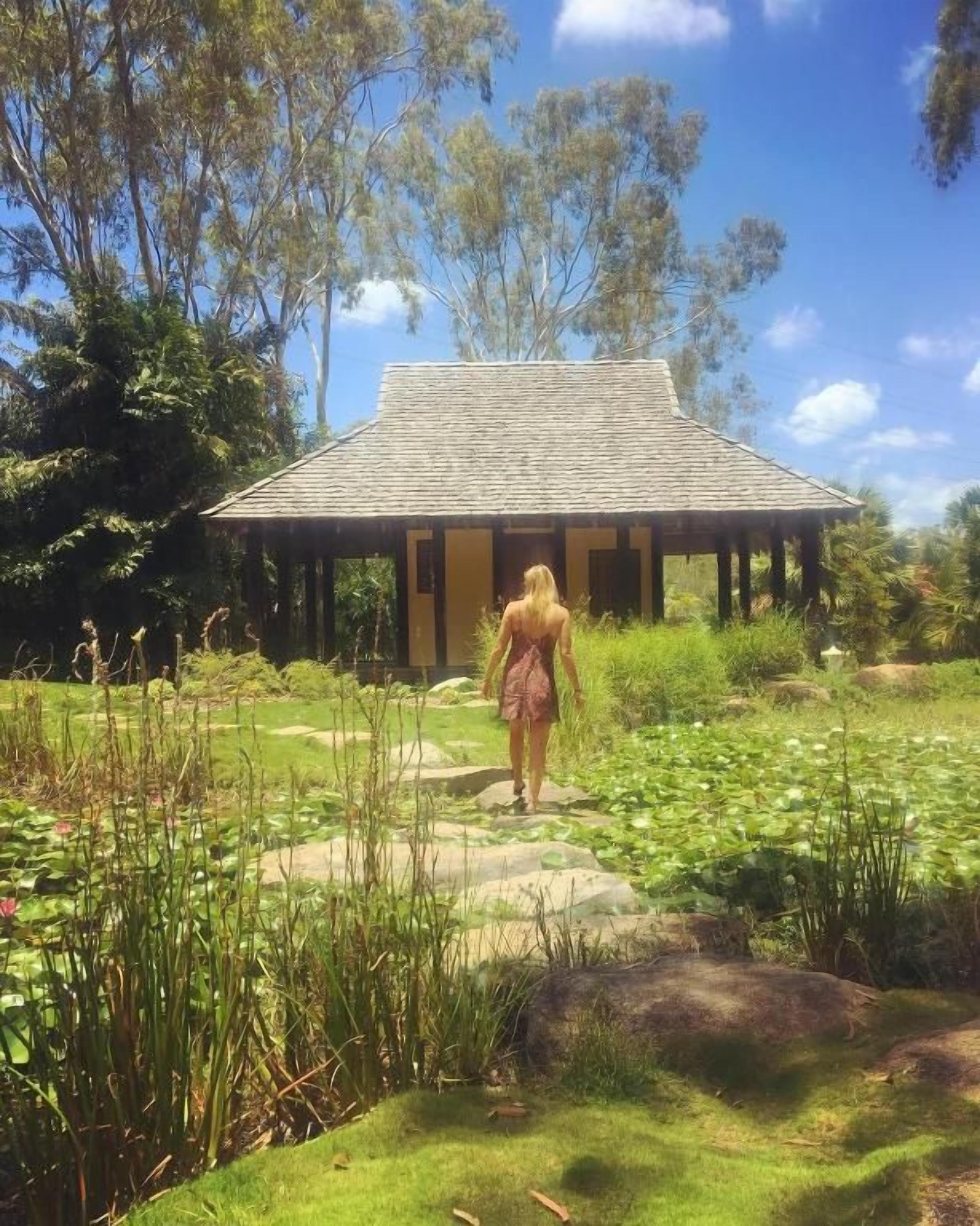 A person walking towards a building with a tiled roof, surrounded by lush greenery and a pond with lily pads.