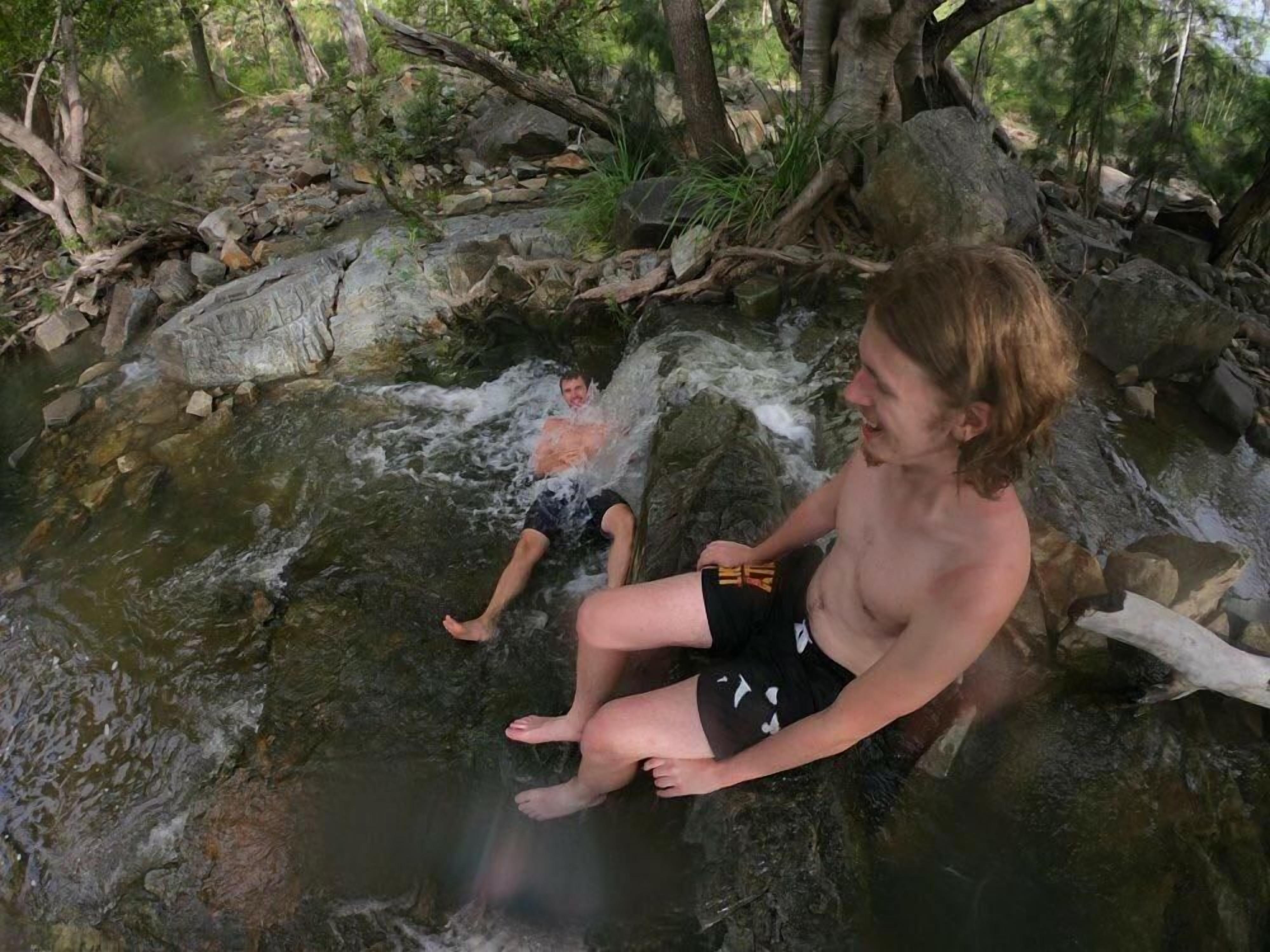 A person sitting on a rock by a flowing stream in a forested area.