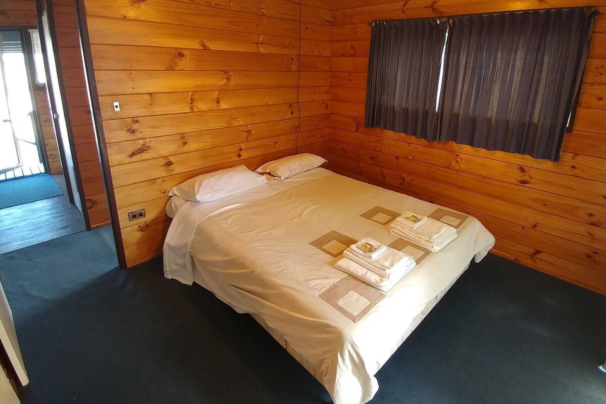 Um quarto com painéis de madeira, uma cama, uma janela com cortinas e uma porta que dá para outro cômodo.