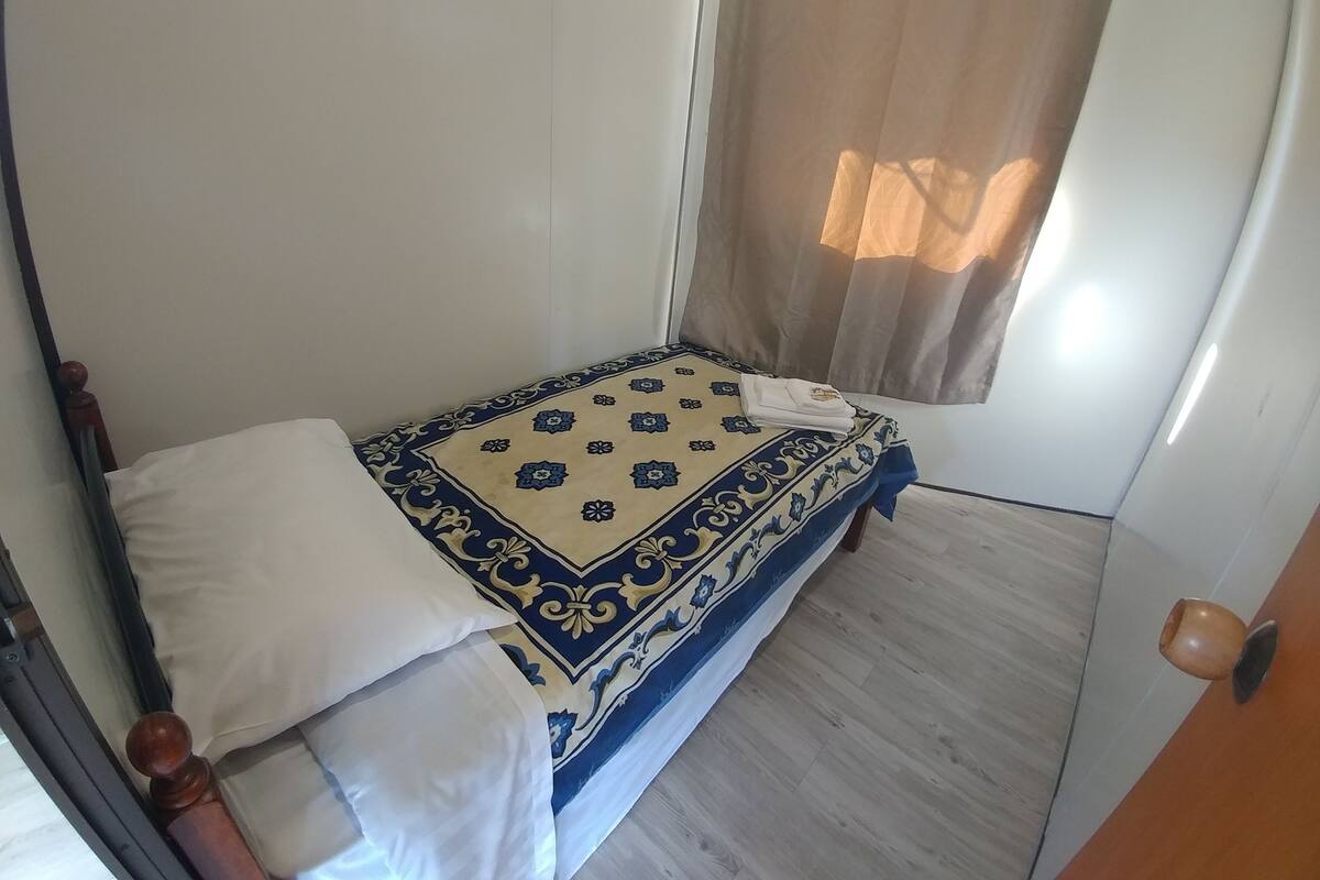 Quarto pequeno com uma cama de solteiro, colcha estampada, janela com cortina e porta de madeira.