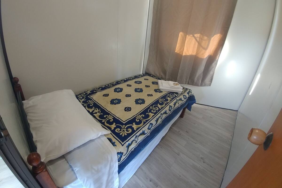 Quarto pequeno com uma cama de solteiro, colcha estampada, janela com cortina e porta de madeira.