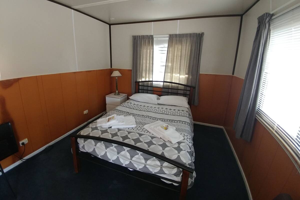 Uma cama de solteiro com edredom estampado, uma mesinha de cabeceira com luminária e cortinas na janela.