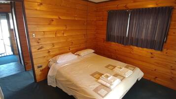 A wooden-paneled room with a bed, a window with curtains, and a door leading to another room.