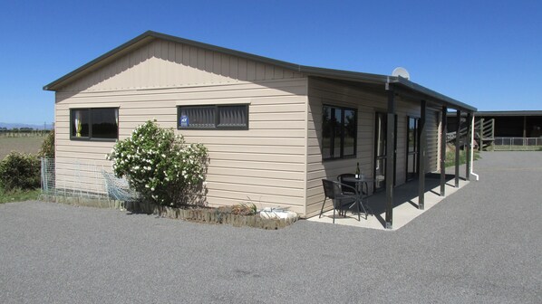 A small, single-story building with a beige exterior, a small porch with a table and chairs, and a bush with white flowers in front.