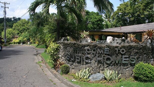 A stone wall with "WELCOME TO SURI HOMES" sign, surrounded by lush greenery and a residential building. - Vacation Villas at Subic Homes (Morong)