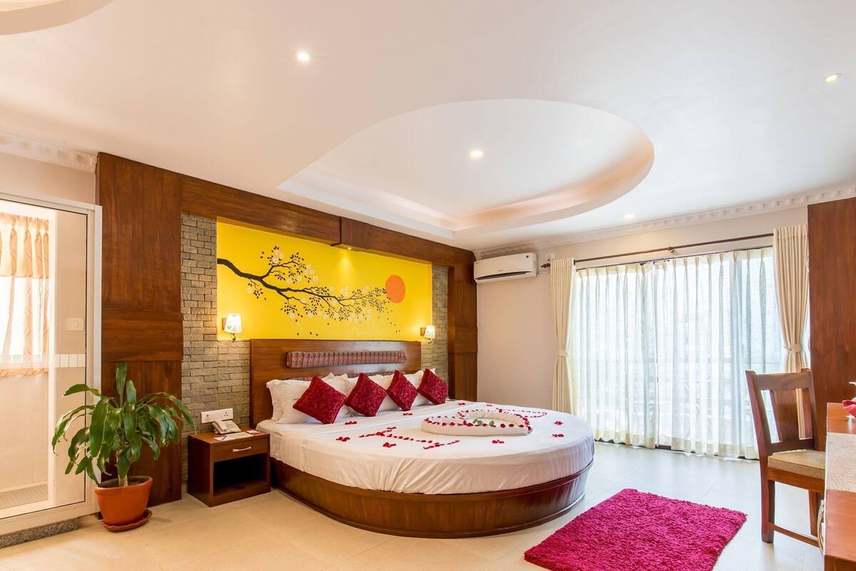 A hotel room with a round bed, a potted plant, a wooden headboard, a wall mural, and a large window with curtains.