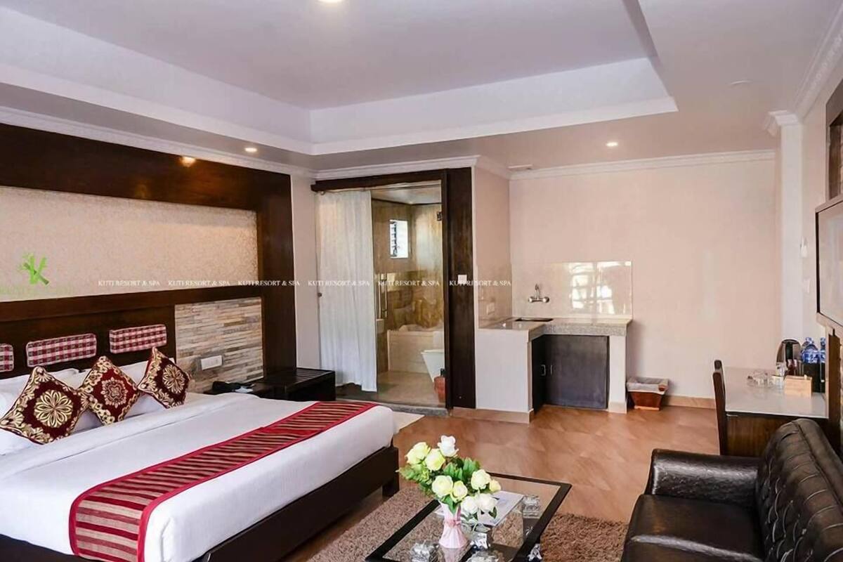 A hotel room with a bed, a coffee table with flowers, a sofa, a desk, and a bathroom visible through an open door.