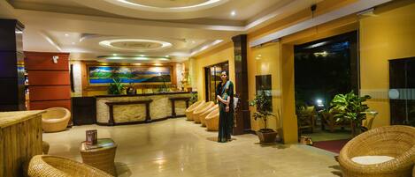 A hotel lobby with a reception desk, wicker furniture, and a large painting on the wall.