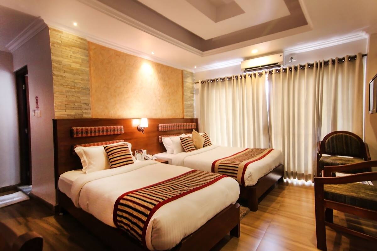 A hotel room with two beds, wooden headboards, a wooden floor, a chair, and a window with curtains.