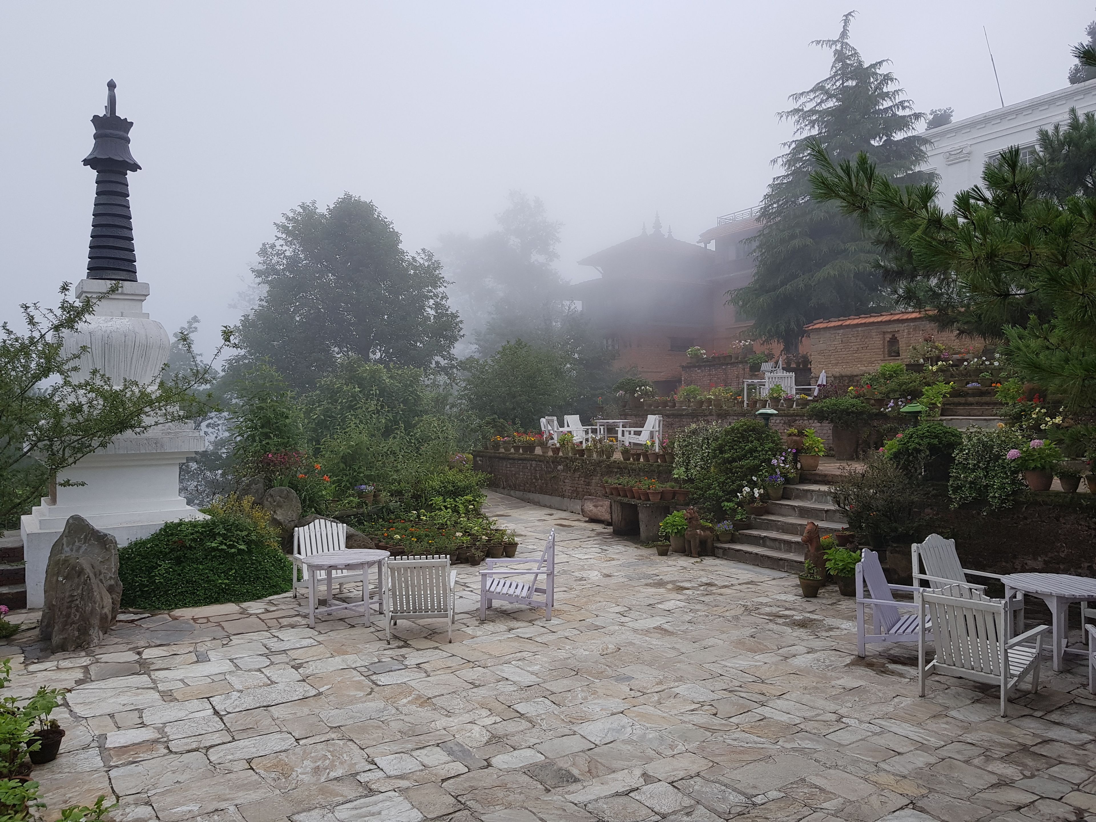 A stone-paved courtyard with white wooden chairs and tables, surrounded by lush greenery and a white pagoda.