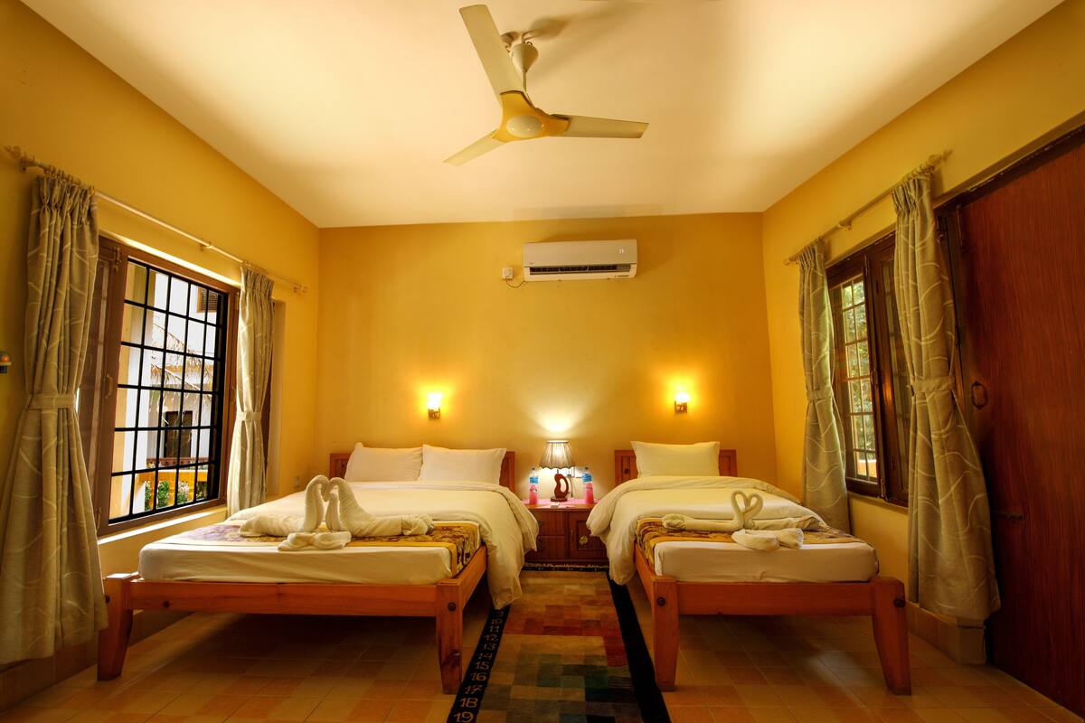A hotel room with two single beds, a ceiling fan, an air conditioning unit, and a window with curtains.