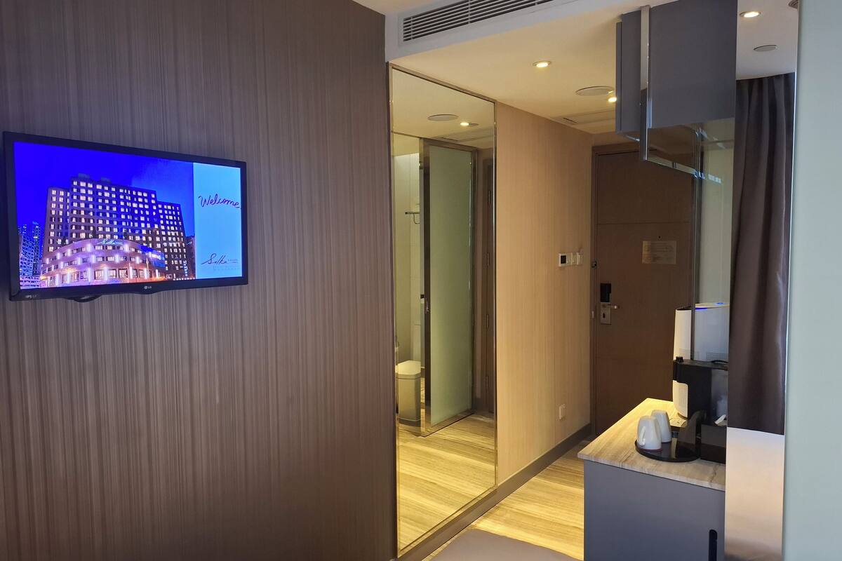 Ein Hotelzimmer mit Fernseher, der eine Stadtansicht zeigt, einem Spiegel, Holzboden, einem kleinen Schreibtisch mit einer Tasse und einer Tür zu einem anderen Raum.
