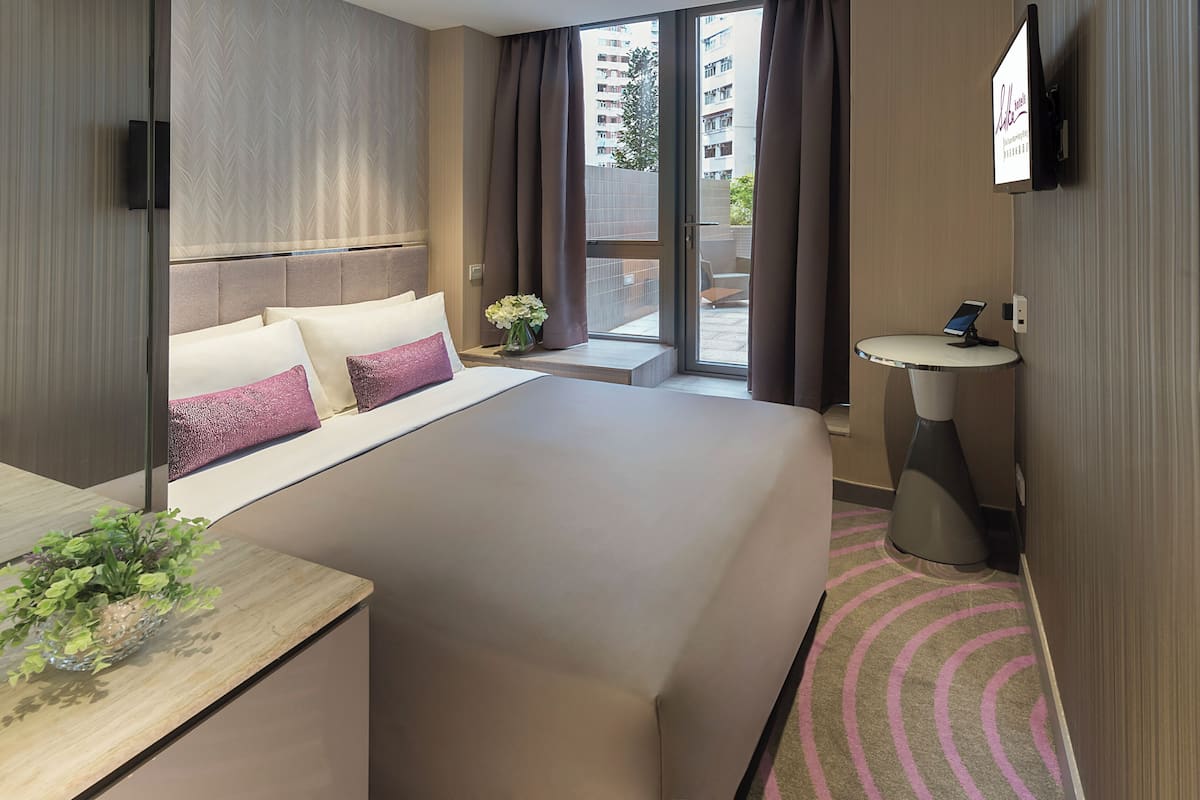 Une chambre d’hôtel avec un grand lit, une table de chevet ornée d’un vase de fleurs, une petite table ronde, un téléviseur fixé au mur et une vue sur l’extérieur.