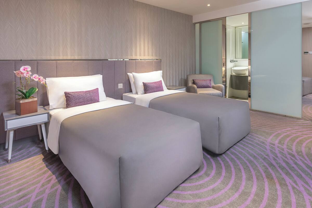 Ein modernes Hotelzimmer mit zwei Betten, einem Schreibtisch, einem Stuhl und einem durch eine Glastür einsehbaren Badezimmer.