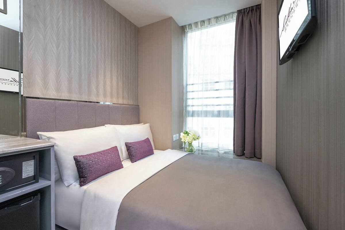 Ein Hotelzimmer mit einem großen Bett, einem Nachttisch, einem an der Wand befestigten Fernseher und einem Fenster mit Vorhängen.