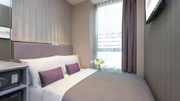 A hotel room with a large bed, a bedside table, a TV mounted on the wall, and a window with curtains.