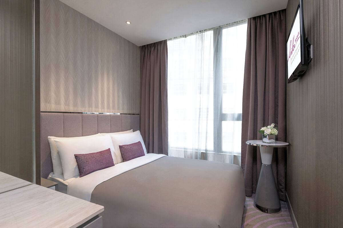 Ein Hotelzimmer mit einem großen Bett, einem kleinen Tisch mit Blumenvase und einem flachbildfernseher an der Wand.