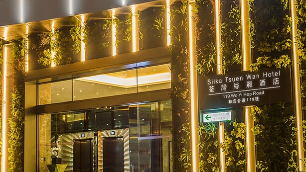 Het uiterlijk van het Silka Tsuen Wan Hotel, met een moderne glazen gevel, een verticale tuin en verlichte signage.