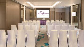 A conference room with rows of white chairs, a stage with a presentation screen, and a carpet with a geometric pattern.