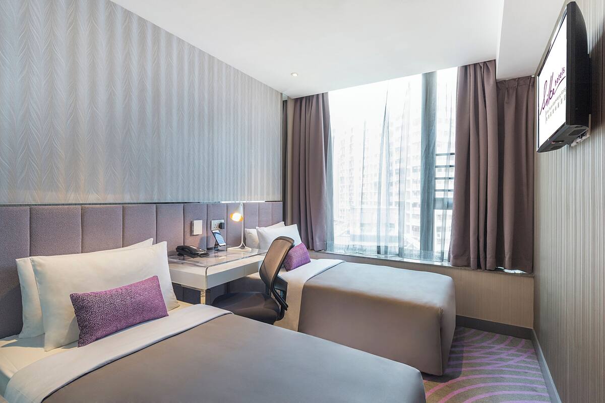 Ein Hotelzimmer mit zwei Betten, einem Schreibtisch mit Lampe, einem Sessel und einem an der Wand befestigten Fernseher.