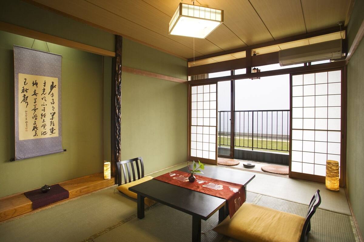 A traditional Japanese room with tatami flooring, a low table, a cushioned bench, a sliding door with shoji screens, and a hanging scroll with calligraphy.