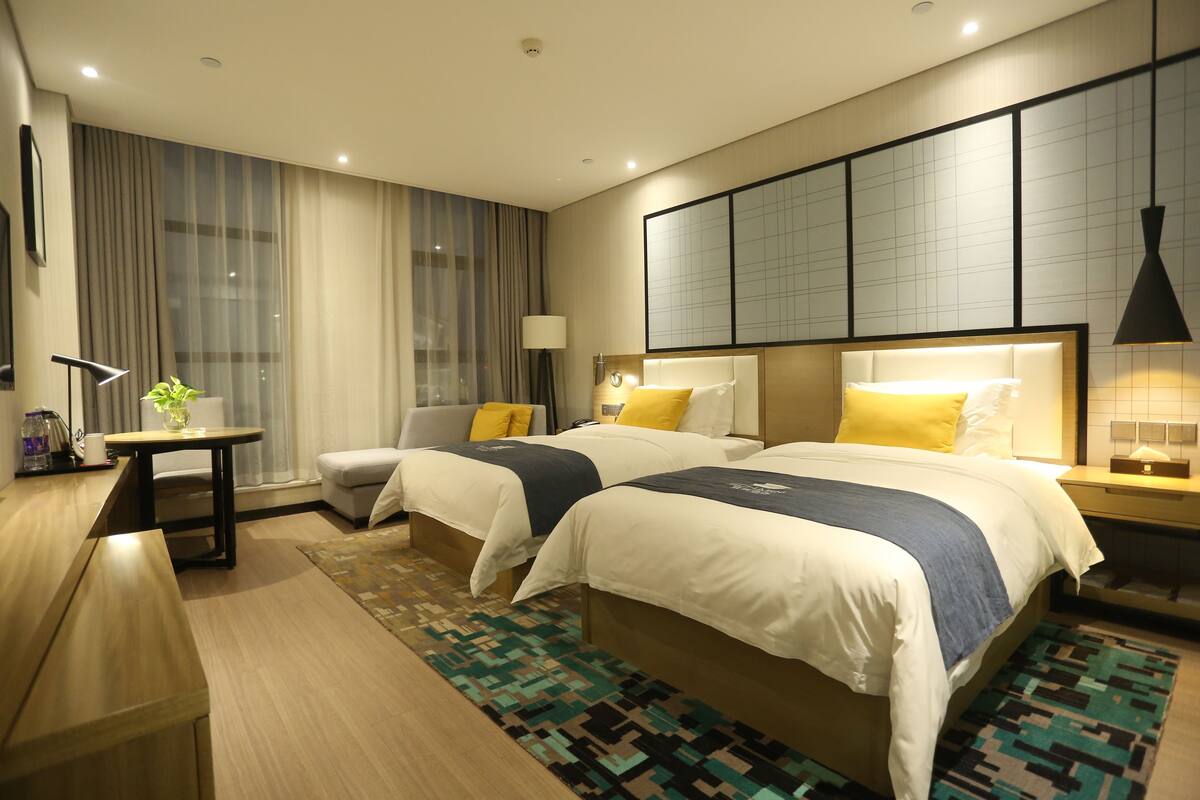 Hotelkamer met twee bedden, een bureau, een stoel, een lamp en een groot raam met gordijnen.