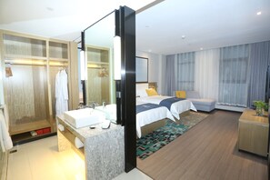 Quarto de hotel moderno com uma cama grande, duas mesinhas de cabeceira, banheiro com pia e espelho, e um closet espaçoso.