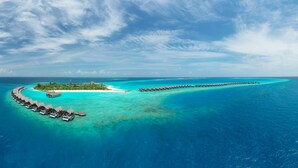 An overwater bungalow resort with a crescent-shaped island, clear turquoise waters, and a long pier connecting the island to the main land.