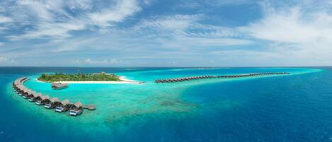 An overwater bungalow resort with a crescent-shaped island, clear turquoise waters, and a long pier connecting the island to the main land.