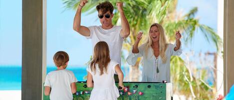 A family playing foosball outdoors with a beach and palm trees in the background.