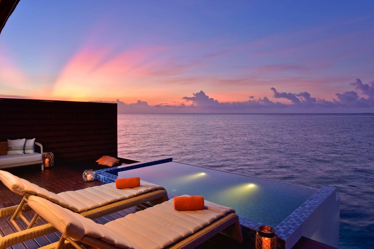 A sunset view from an infinity pool with lounge chairs and a wooden deck.