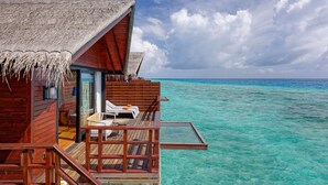 A wooden overwater bungalow with a thatched roof, a glass door leading to a lounge area with white chairs and a small table, and a clear view of the turquoise sea. - Grand Park Kodhipparu (Kuda Vattaru)