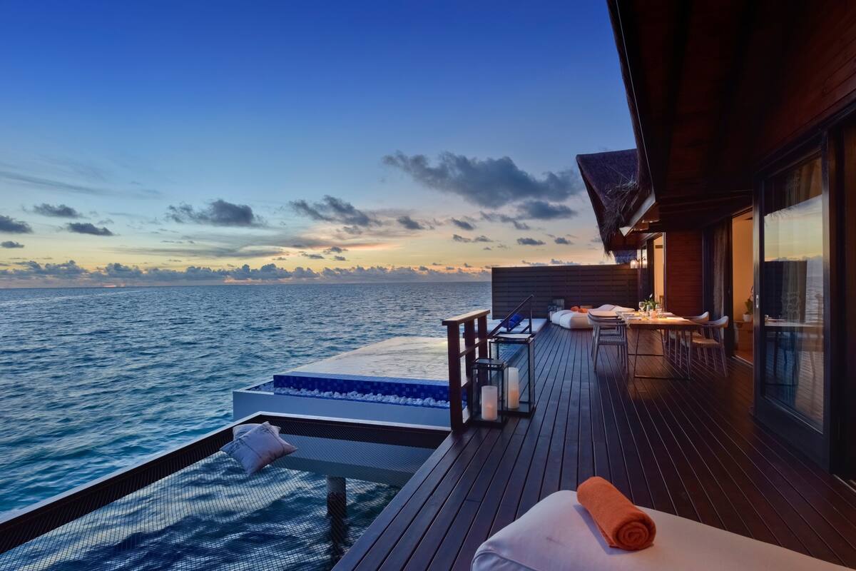 A wooden deck with a view of the ocean, a table with chairs, and a small seating area with cushions.