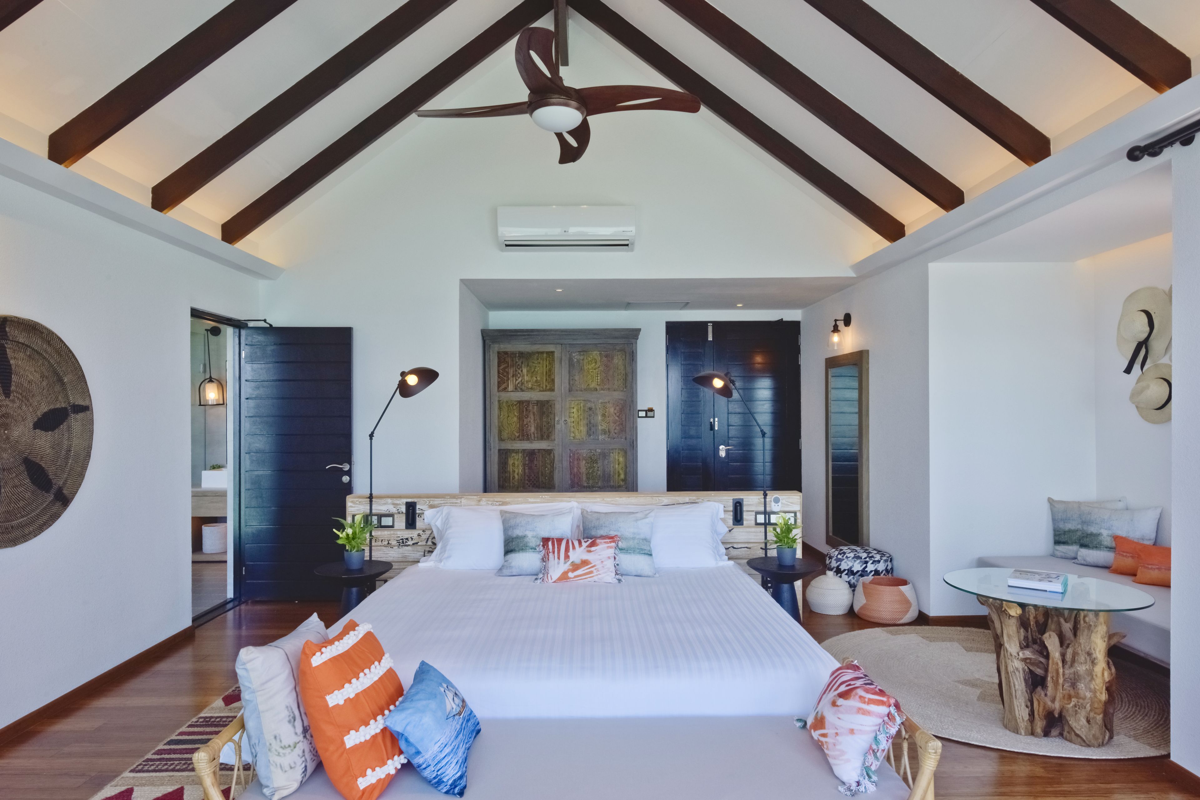 Une chambre spacieuse avec un grand lit, des poutres apparentes, un ventilateur de plafond et une vue sur le paysage urbain, visible à travers une grande fenêtre.