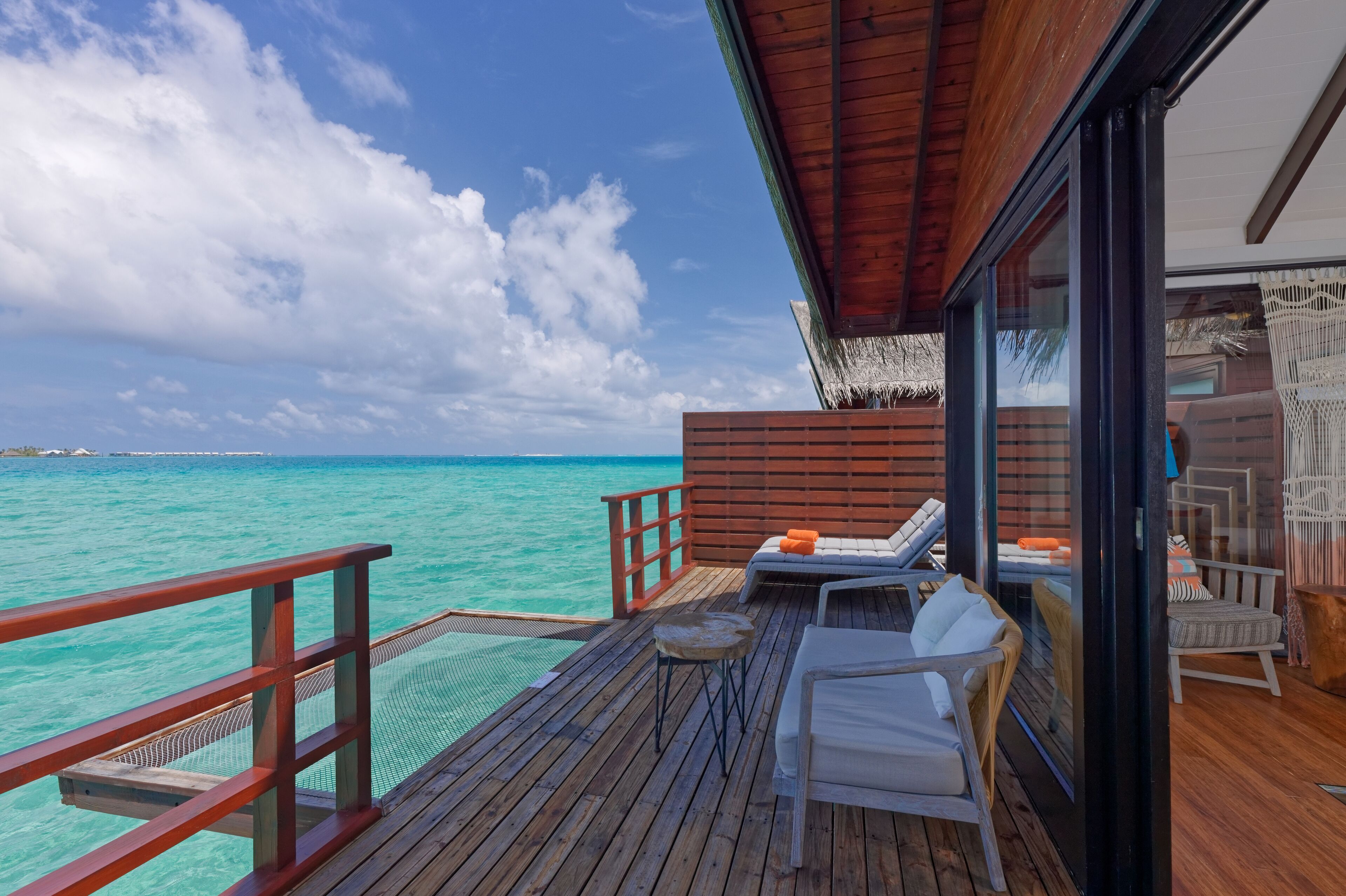 A wooden deck with a view of turquoise water, a glass door, and a seating area with cushions.