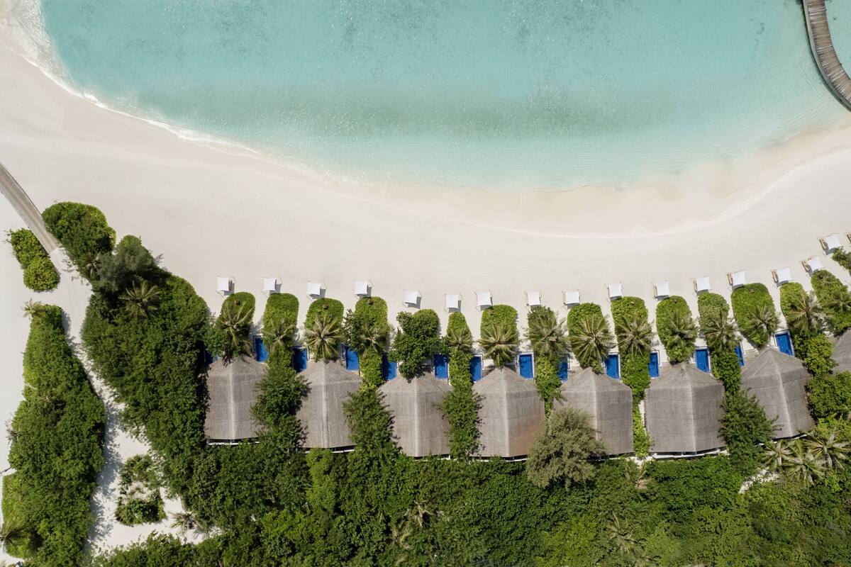 An aerial view of a beach resort with rows of thatched-roof huts, a sandy beach, and a clear blue sea.