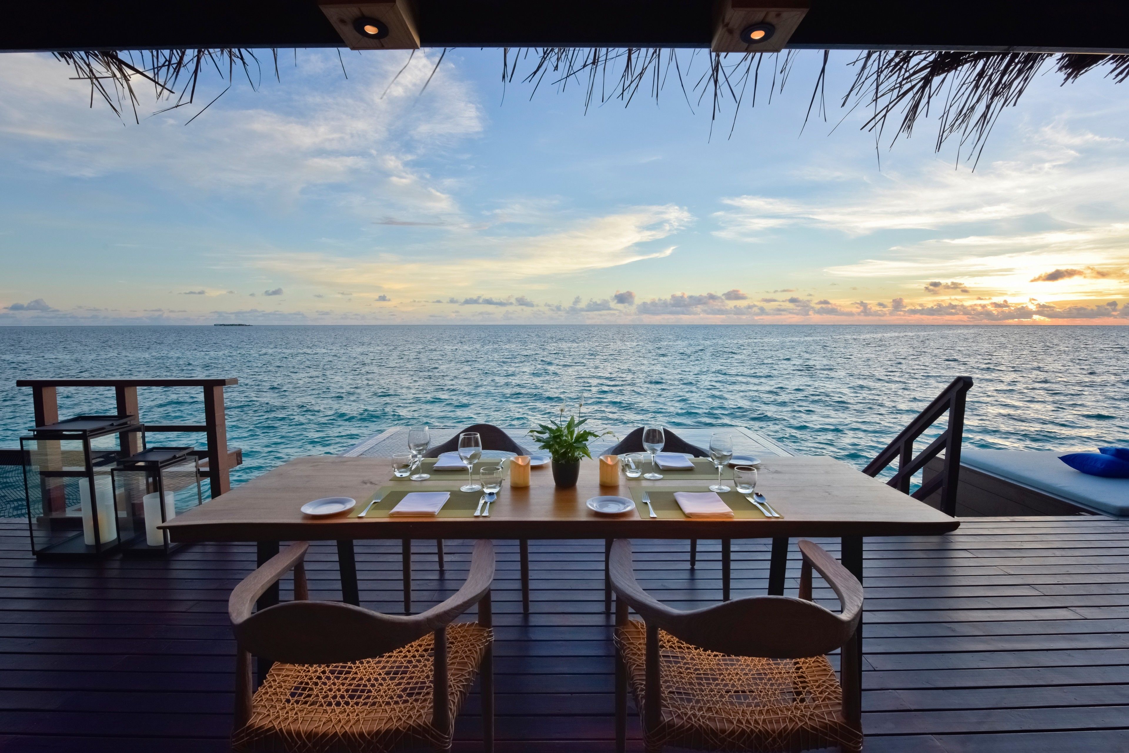 A dining table set on a wooden deck with a view of the ocean and a sunset.
